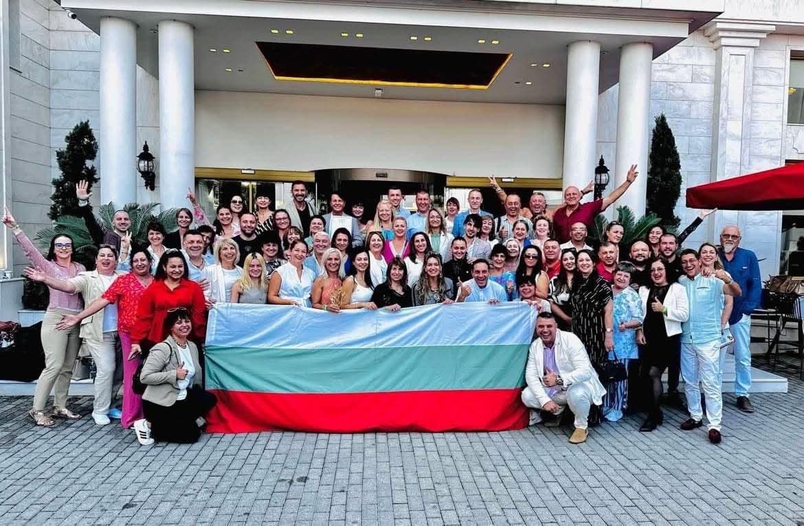 Голяма група партньори от екипа позират усмихнати пред входа на хотел, държейки българското знаме.