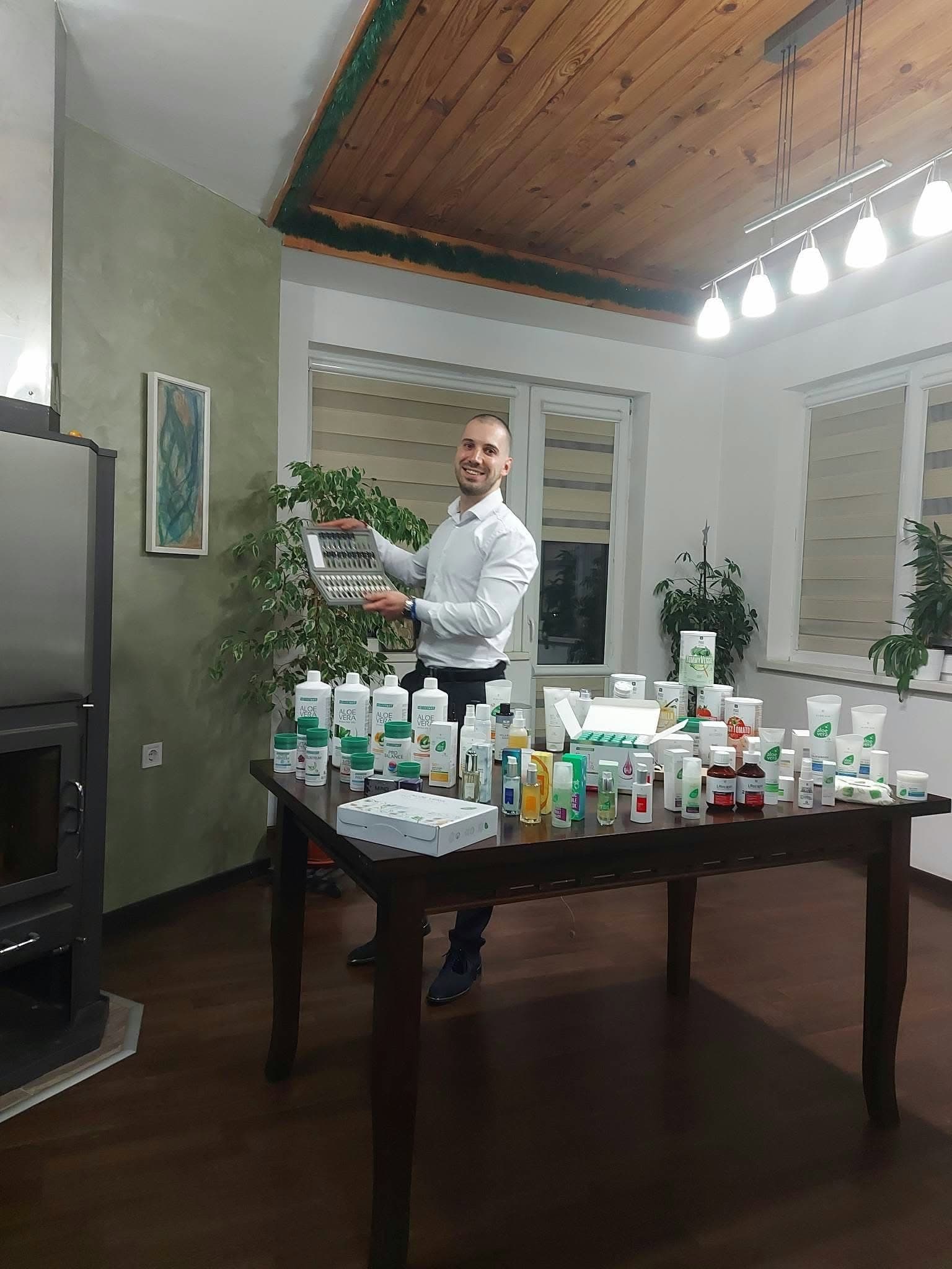 Йордан Колев в дома си показва кутия с продукти на LR, пред него маса, подредена с напитки с алое вера, добавки и козметика.
